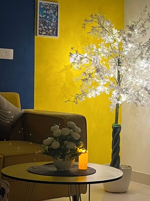 Ein Zimmer mit gelber Wand, einem braunen Sofa, einem runden Tisch mit Kerze und einer Topfpflanze mit weißen Blüten.