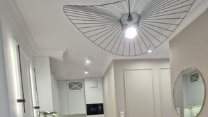 A modern living room with a grey sofa, a dining area with a white table and chairs, a kitchen in the background, and a unique ceiling light fixture.