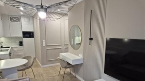 A modern living room with a flat-screen TV, a dining area with a round table and chairs, a sofa, and a kitchen in the background.