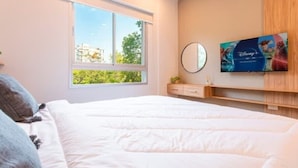 Une chambre avec un grand lit, une fenêtre, un téléviseur fixé au mur et une petite commode.