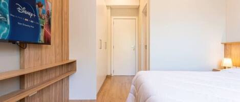 A hotel room with a bed, a TV mounted on the wall, a wooden wardrobe, and a door leading to another room.