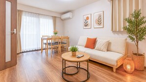 Ein modernes Wohnzimmer mit Sofa, einem Esstisch mit Stühlen, einem kleinen runden Tisch mit Fernbedienung, einer Zimmerpflanze und einer Wand-Klimaanlage.