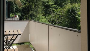 Un balcón con mesa y sillas, una pared blanca y vistas a árboles.