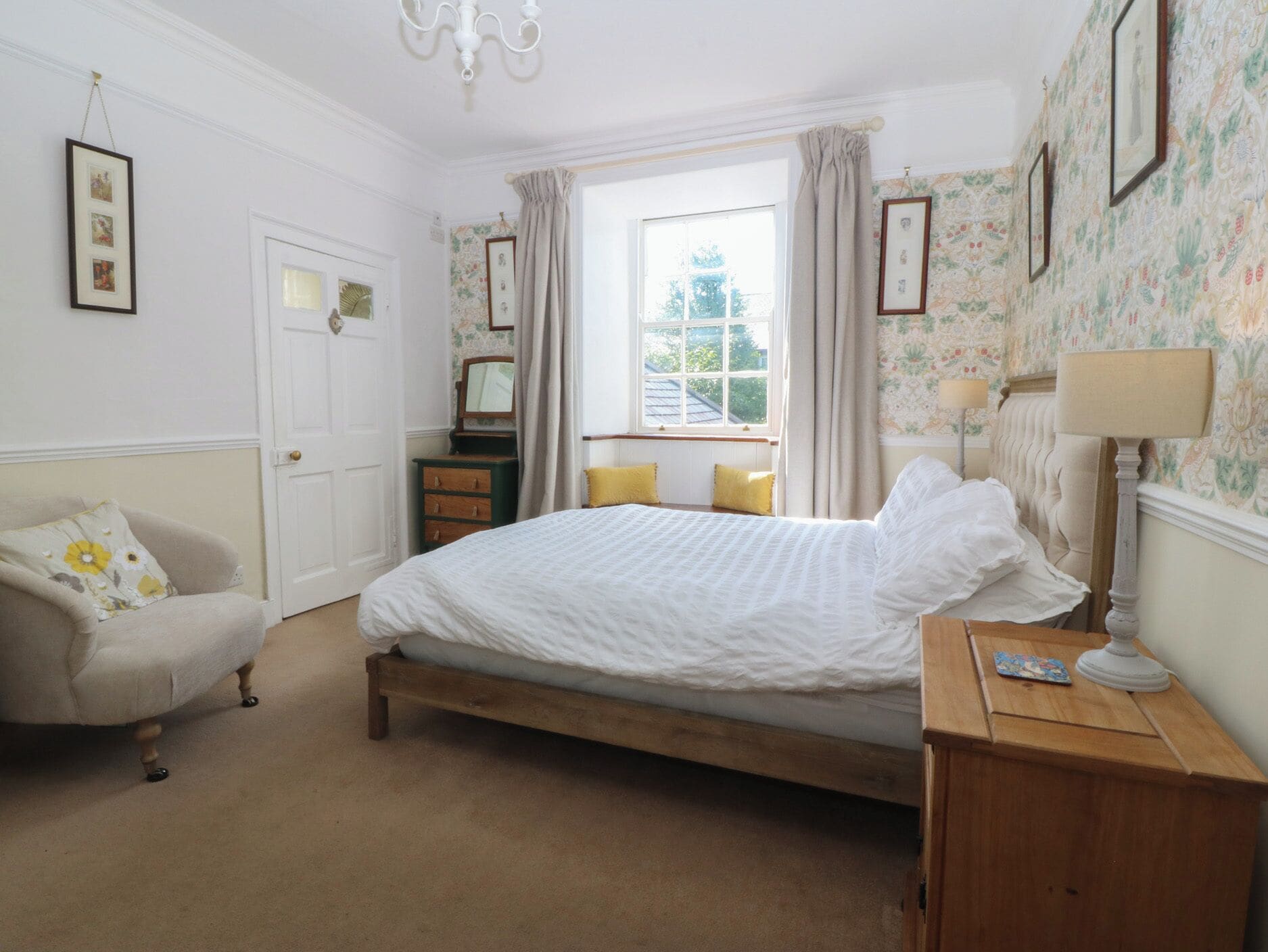A bedroom with a bed, a dresser, a chair, a window with curtains, and a chandelier.