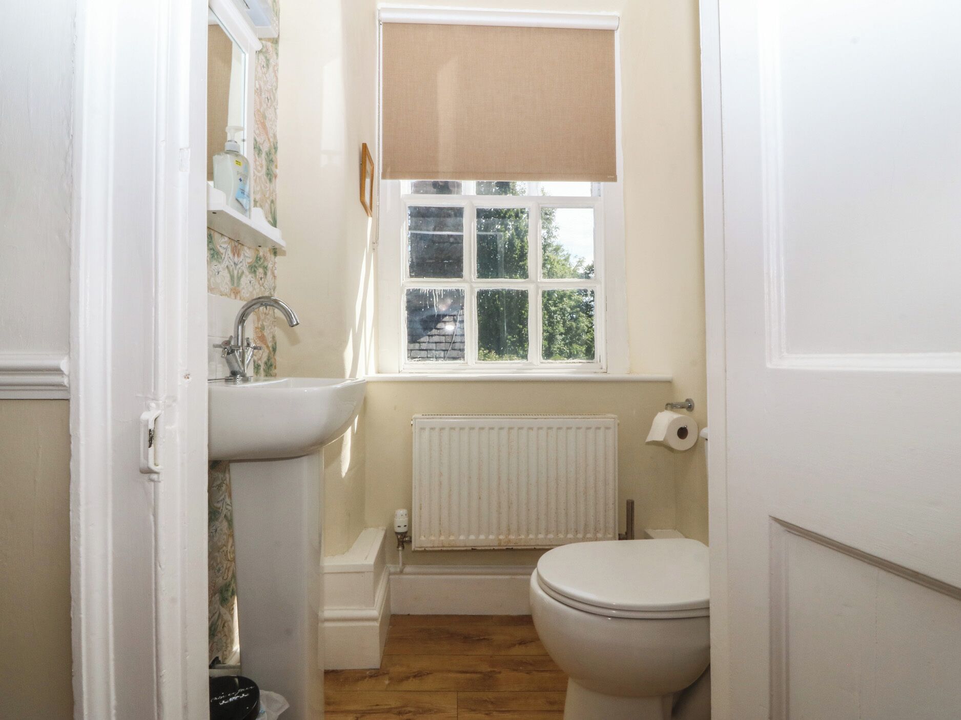 Un baño pequeño con un lavamanos blanco, un inodoro, una ventana con persiana color beige y un radiador.