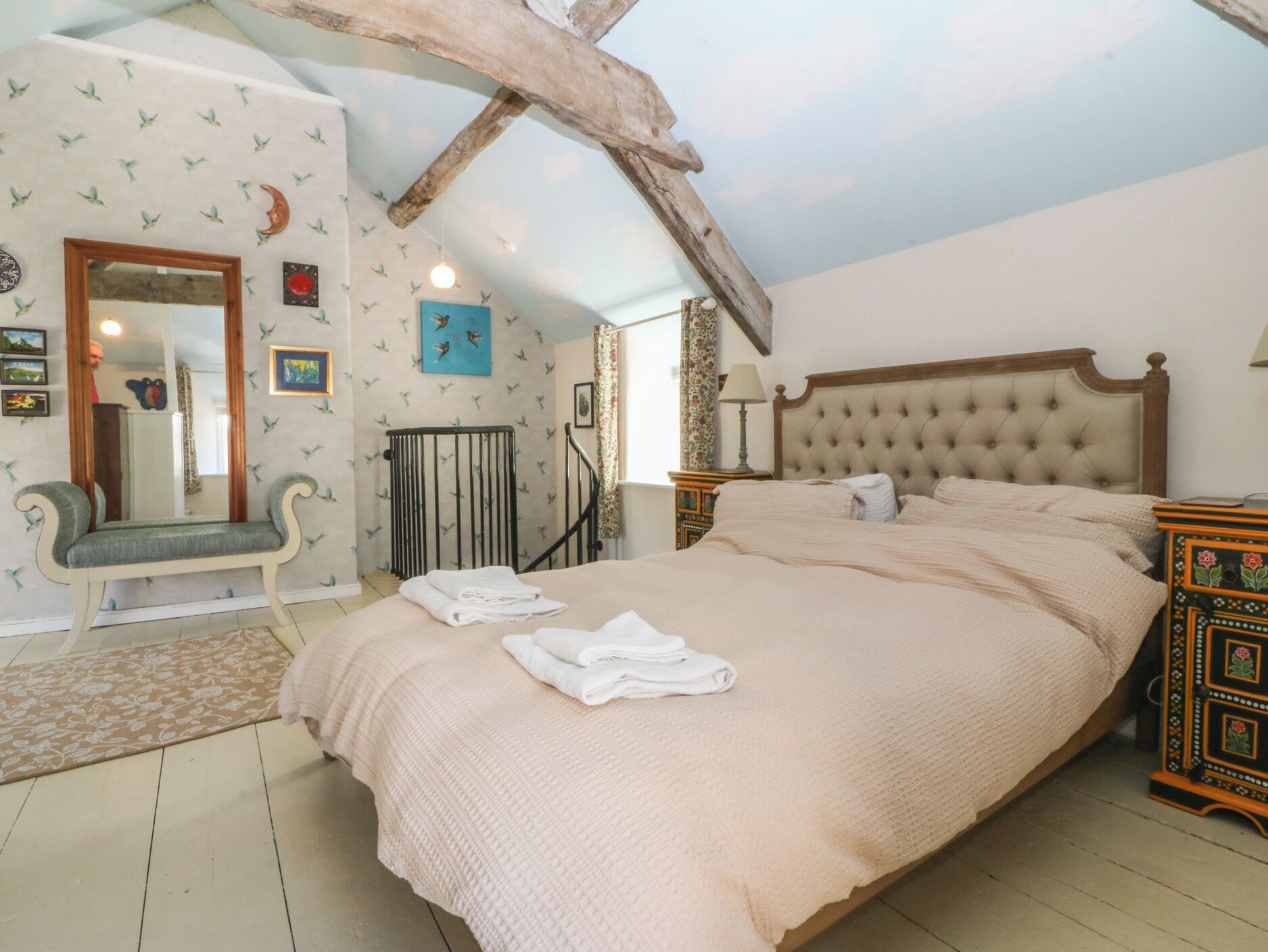 Un dormitorio con una cama grande, cabecera de madera, un banco, un espejo y vistas al cielo a través de una ventana.