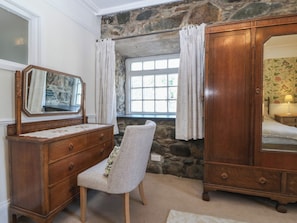 Una habitación con pared de piedra, un aparador de madera con espejo, una silla y una ventana con cortinas.