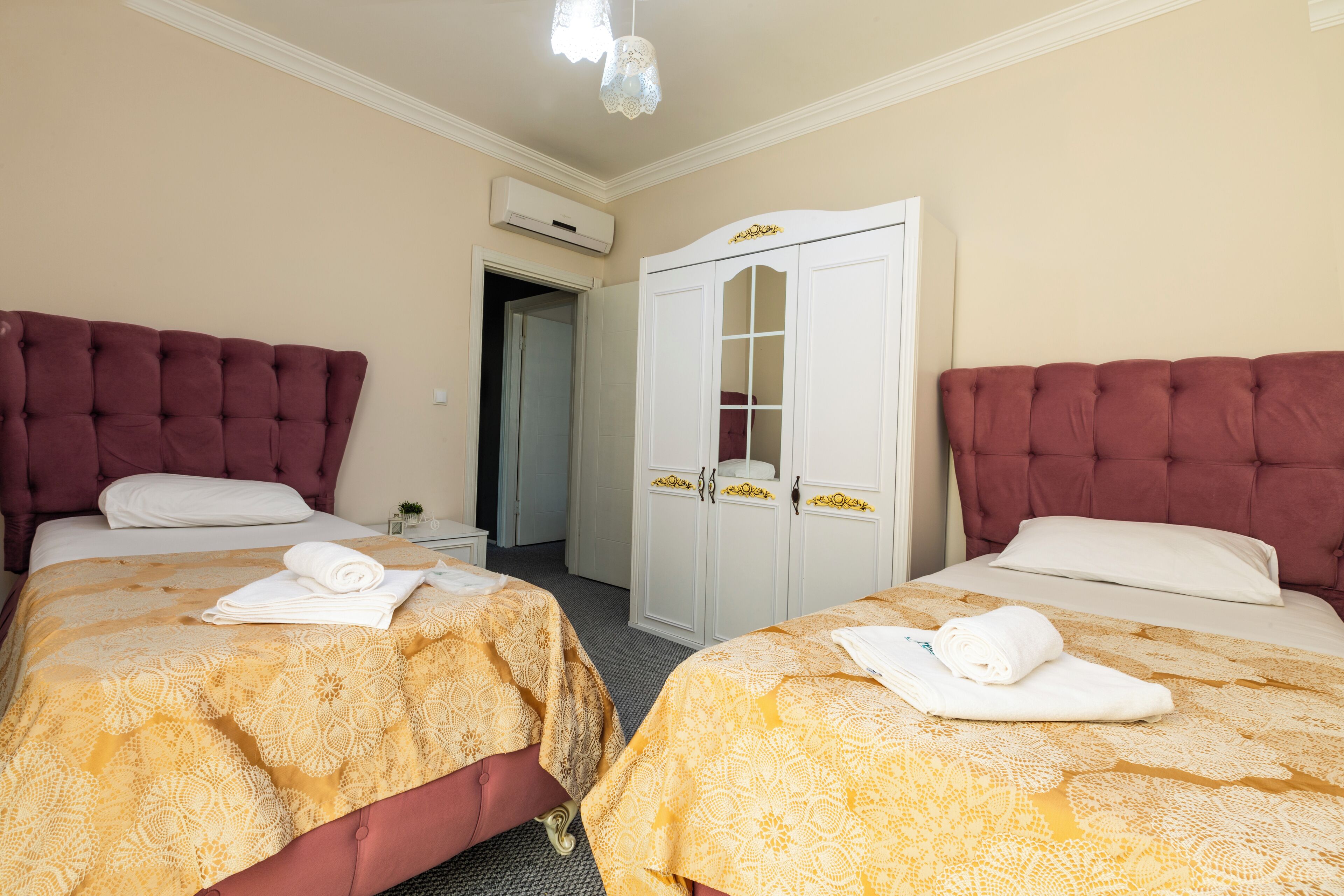 A hotel room with two beds, each with a red headboard and a patterned bedspread. There is a white wardrobe with glass doors and a small table with a lamp. An air conditioning unit is mounted on the wall.