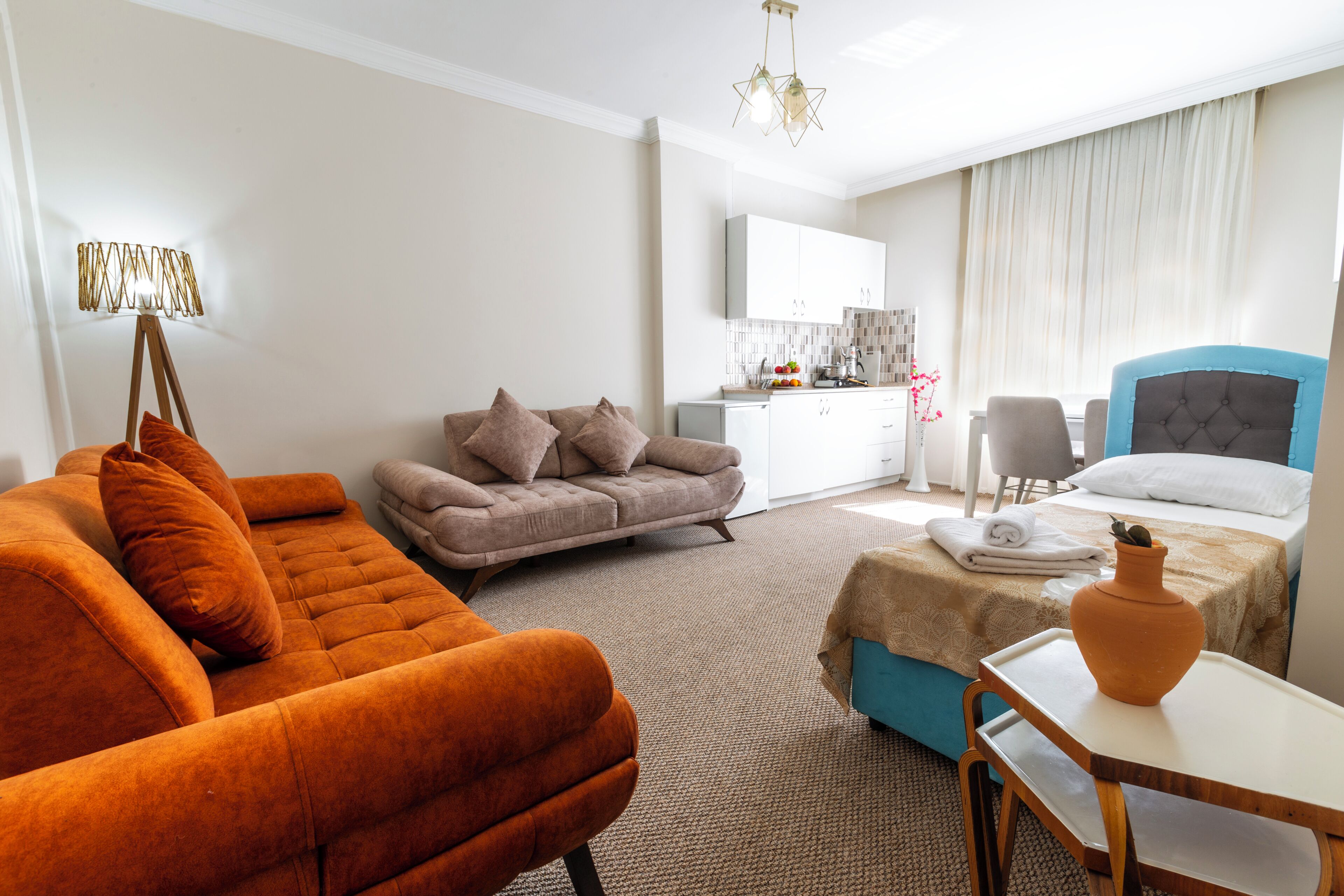 A modern hotel room with a sofa, a chair, a bed, a small table, and a kitchen area in the background.