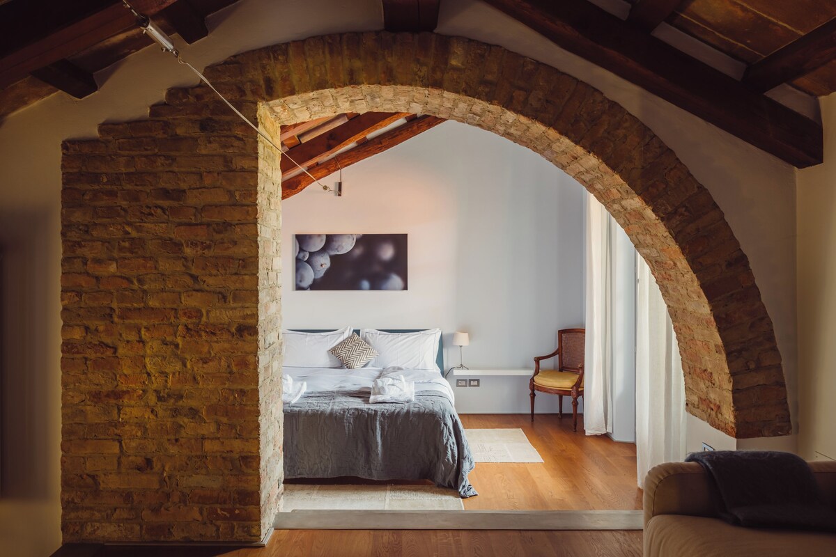 Un dormitorio con un arco de ladrillo, una cama con cubrecama gris, un cuadro abstracto colgado en la pared, suelo de madera y una silla.