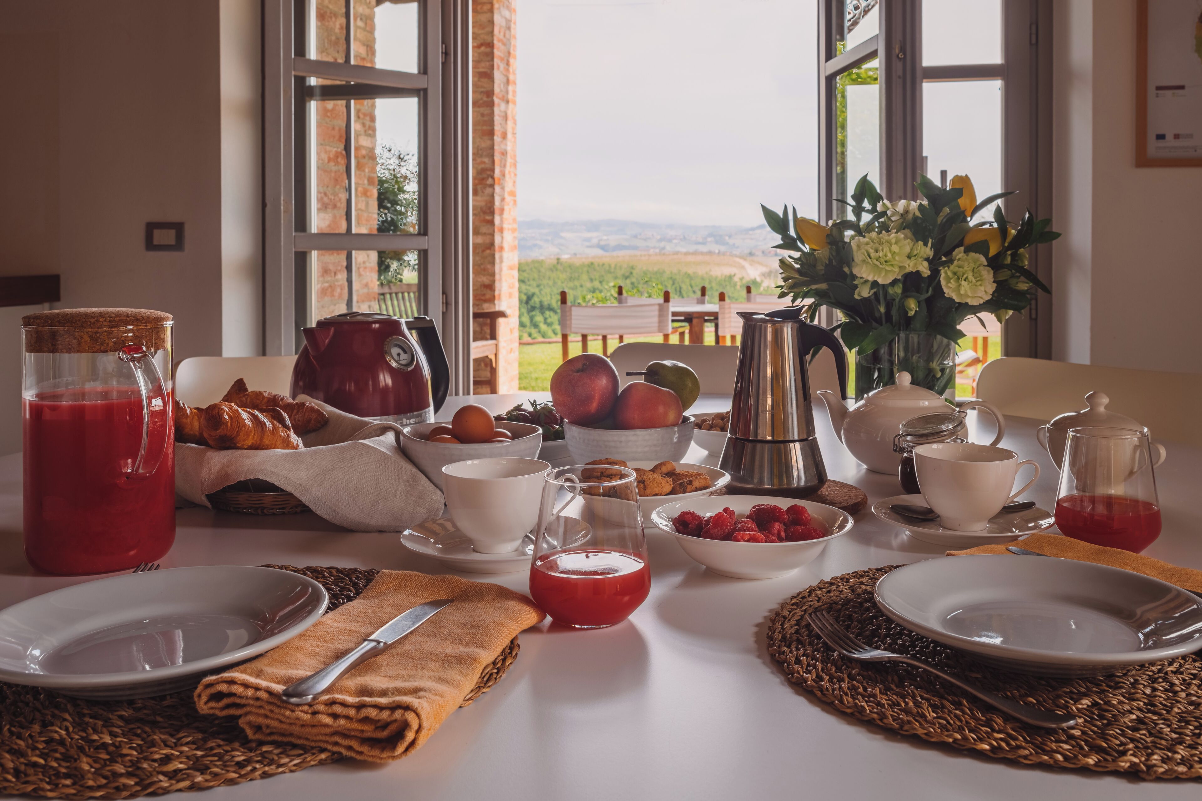 A breakfast table with a view of the countryside, including a basket of croissants, bowls of fruit, and a vase of flowers.