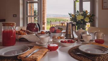 A breakfast table with a view of the countryside, including a basket of croissants, bowls of fruit, and a vase of flowers.