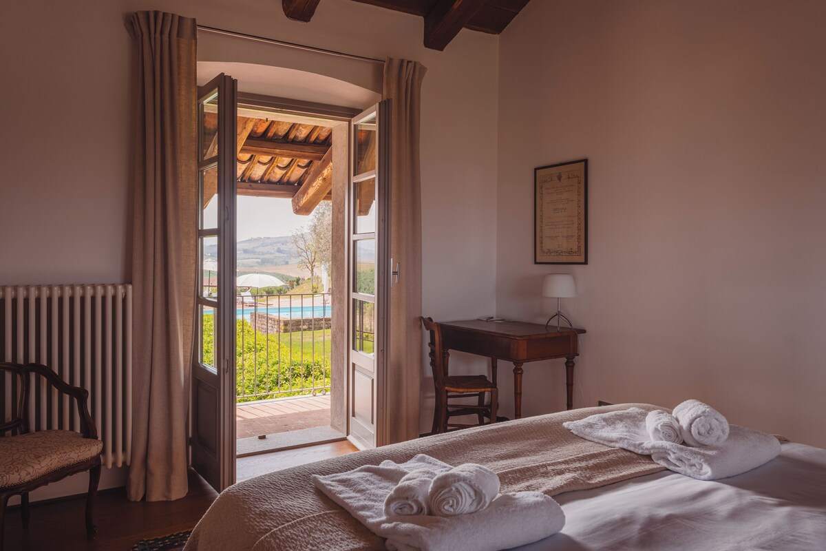 Habitación de hotel con cama, una silla, un escritorio, una lámpara y vistas a una piscina y vegetación.