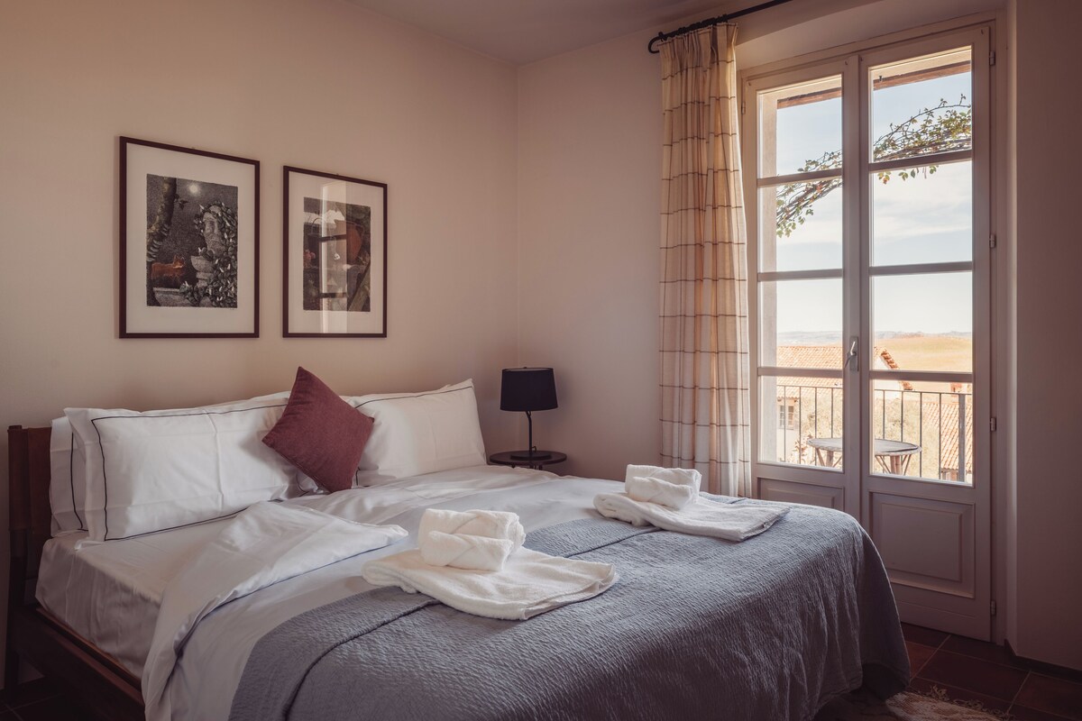 Habitación de hotel con cama, dos cuadros, una mesita de noche con lámpara y un balcón con vistas.