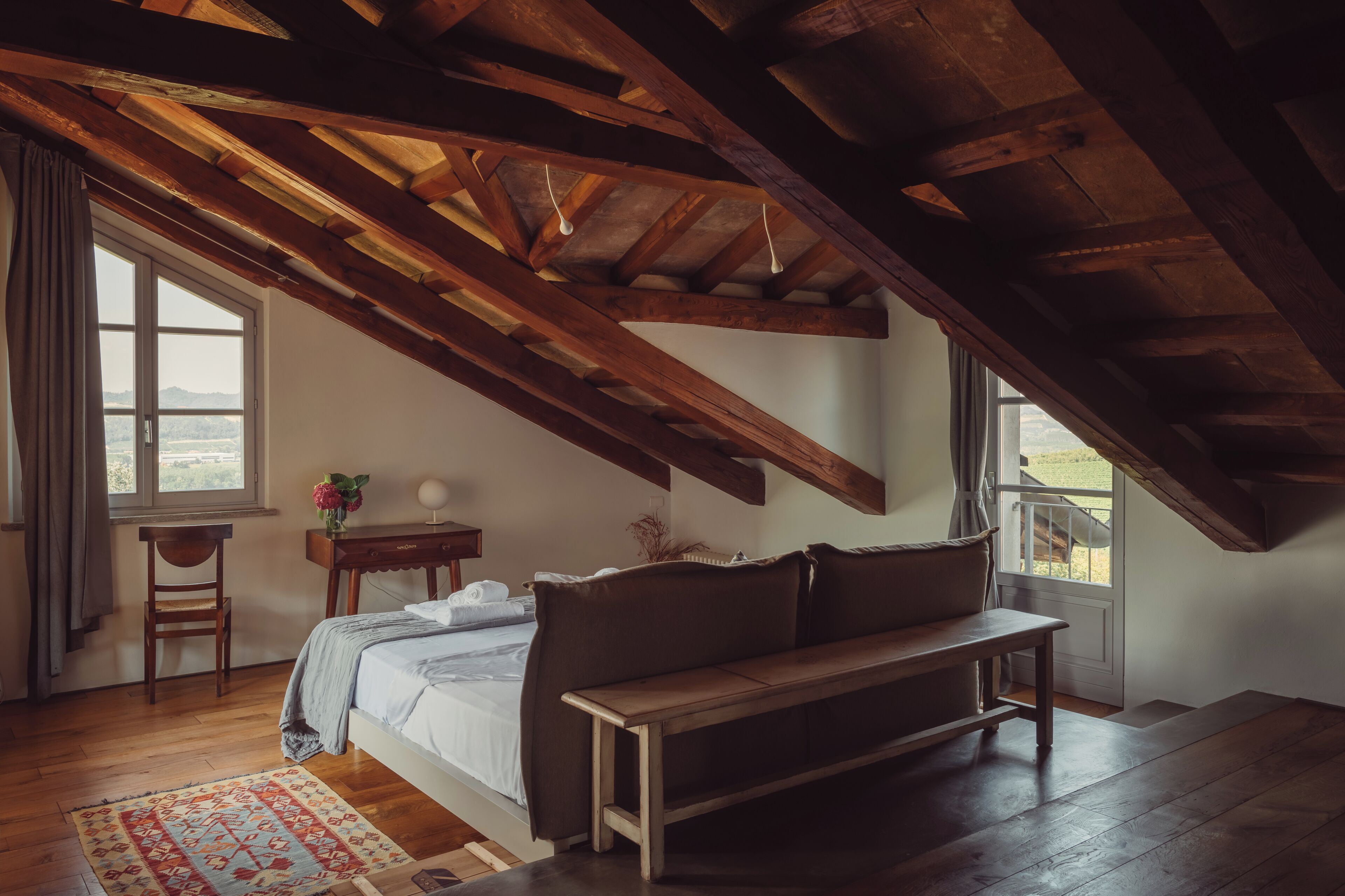 A bedroom with a wooden ceiling, a bed, a bench, a desk, and a window with a view.