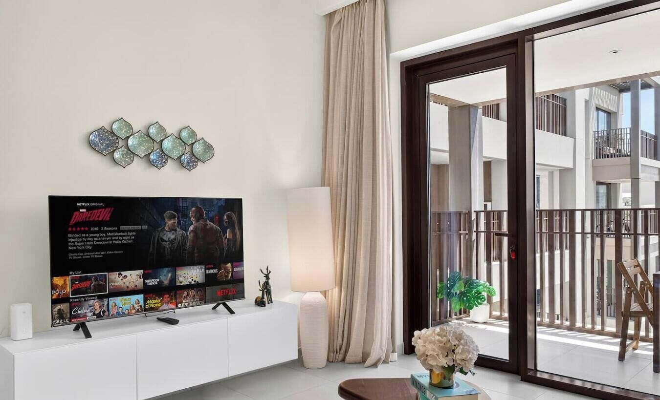 A modern living room with a flat-screen TV, a white entertainment unit, a large window with a view of a balcony, and a decorative wall piece.