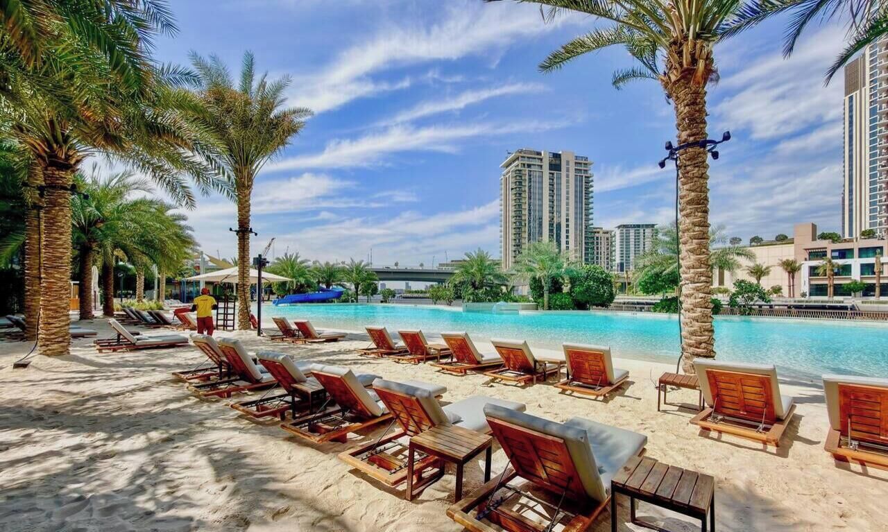 A poolside area with lounge chairs and palm trees, a clear sky, and high-rise buildings in the background.