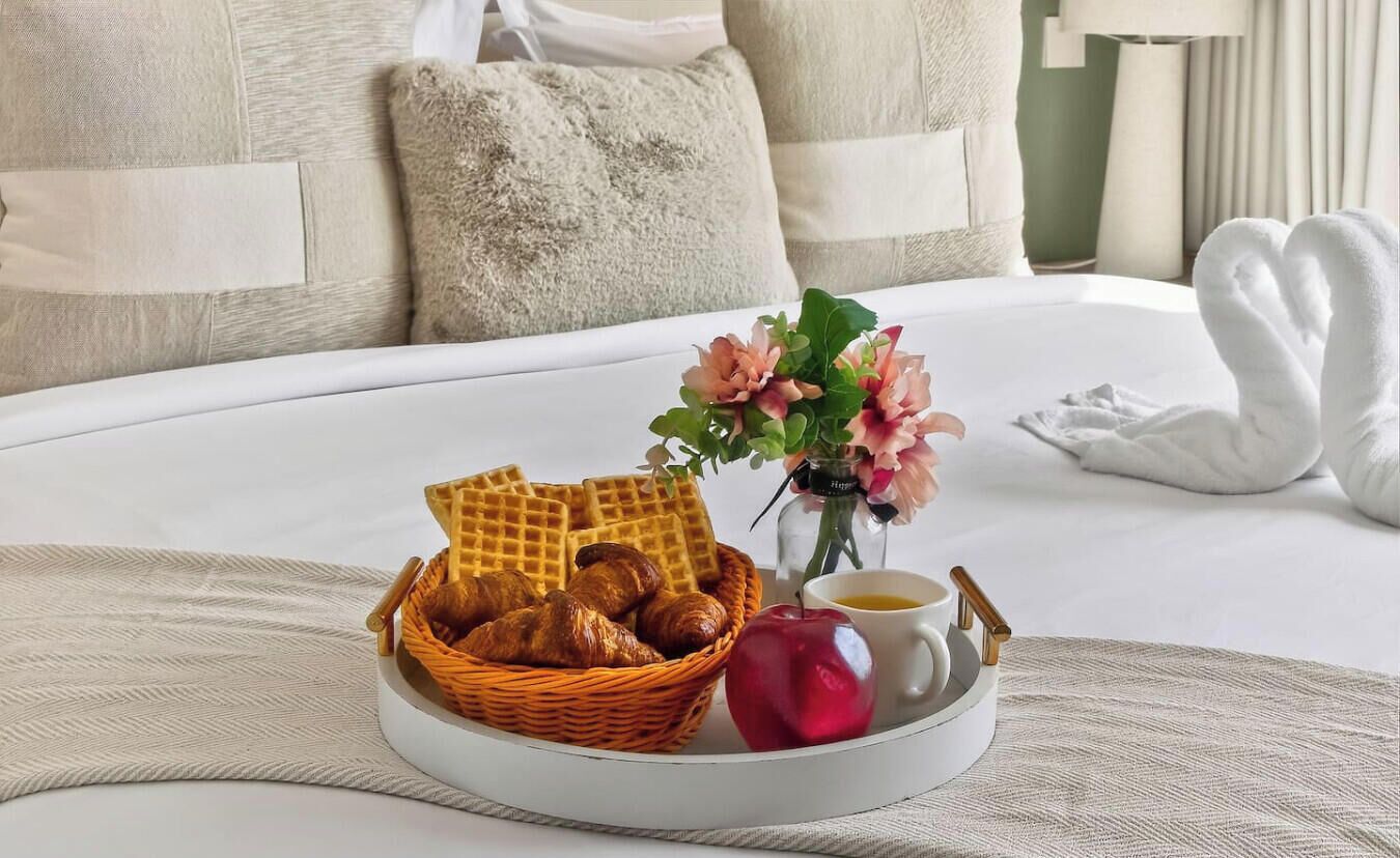 Una cama bien hecha con edredón blanco, una cesta de mimbre con croissants, una manzana roja, una taza de café y un jarrón con flores.