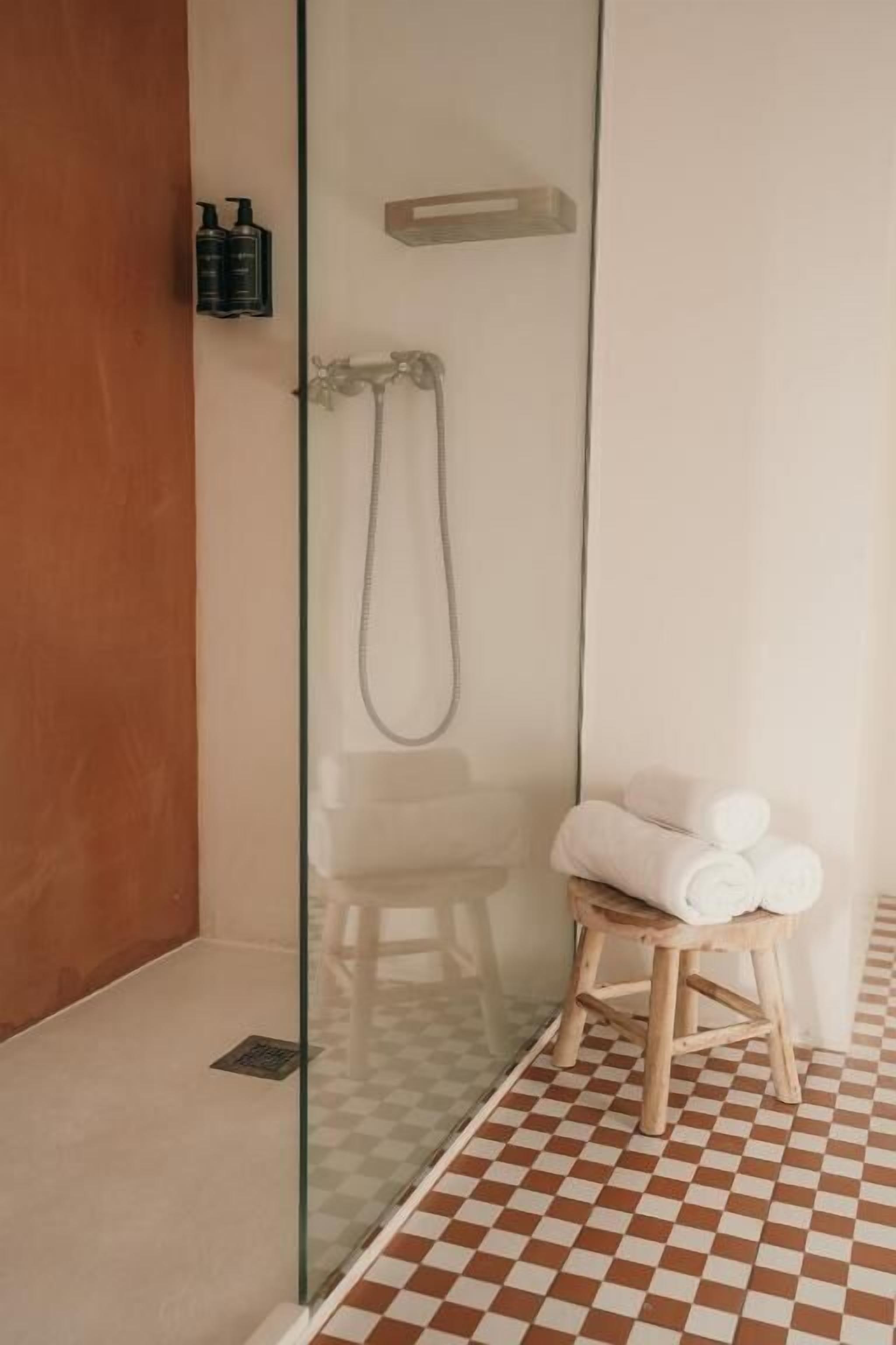 Ducha envuelta en vidrio con cabezal de ducha de mano, un taburete de madera con toallas y un suelo con estampado de cuadros.