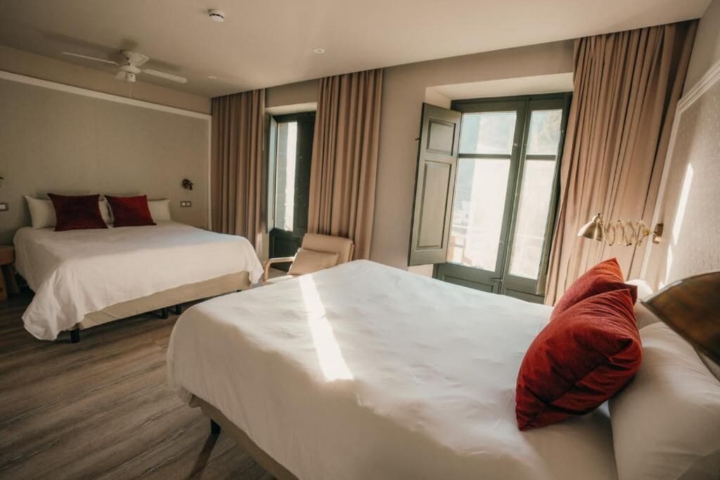 Une chambre d’hôtel avec deux lits, un ventilateur de plafond, de grandes fenêtres avec des rideaux et une vue sur la ville.