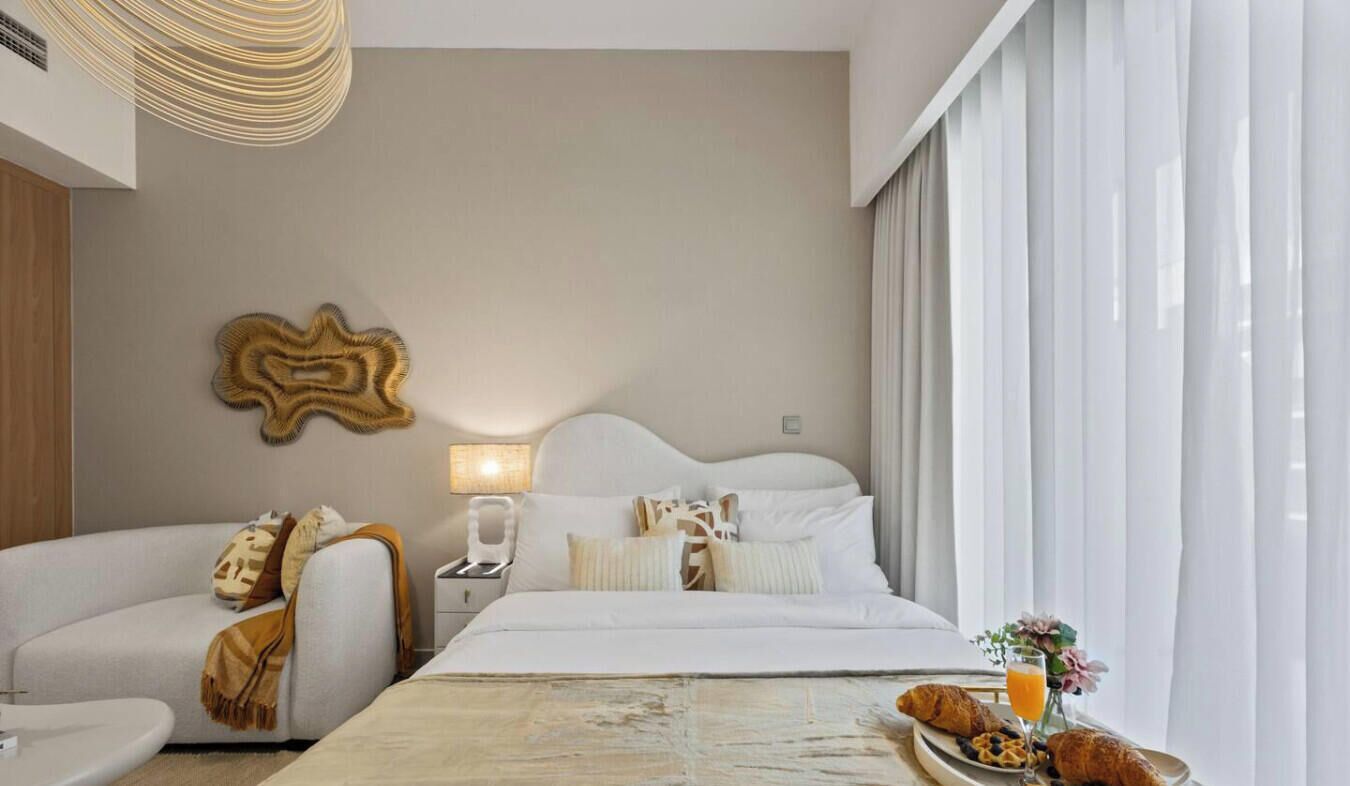 A modern hotel room with a bed, a sofa, a nightstand with a lamp, and a breakfast tray with croissants and juice.