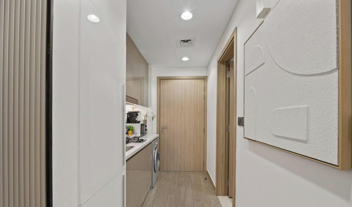 A narrow hallway with a washing machine, a wooden door, and a wall-mounted light fixture.