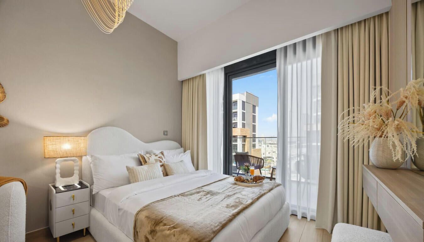 A hotel room with a large bed, a bedside table with a lamp, a view of the city through the window, and a decorative vase on the windowsill.