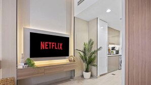Uma sala moderna com televisão de tela plana exibindo 'NETFLIX', uma mesinha de madeira, um vaso de planta e uma cozinha ao fundo.