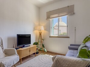 Olohuoneessa on taulutelevisio, puinen kaappi, lattiavalaisin, sohva ja suuri ikkuna, josta on näkymä rakennukseen.