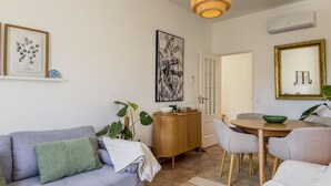 Ett vardagsrum med en soffa, ett matbord, stolar, en hängande lampa, ett skåp och konst på väggen.
