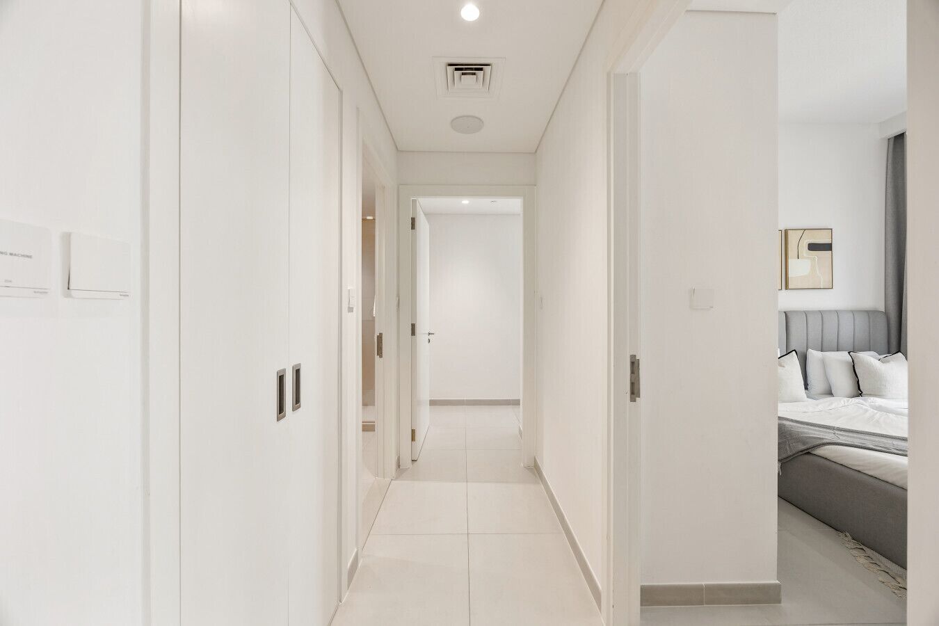 A hotel corridor with white walls, a tiled floor, and a bed with a grey headboard in the adjacent room.