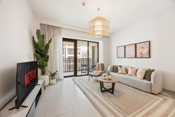 A modern living room with a flat-screen TV, a sofa, a coffee table, a large plant, and a balcony with a railing. - Charming 2 BR in Surf 2 (Dubai)