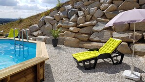 Une piscine avec des marches en bois, deux chaises longues jaunes dotées de coussins, un mur en pierre et un parasol violet.