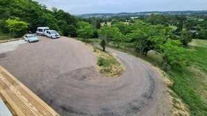 Un parking en gravier où sont stationnés des véhicules, entouré de verdure et offrant une vue sur des collines lointaines.