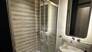 A modern bathroom with a glass-enclosed shower, a marble-tiled wall, a large mirror, and a white sink. - Syntagma Divine Guestrooms (Athens)