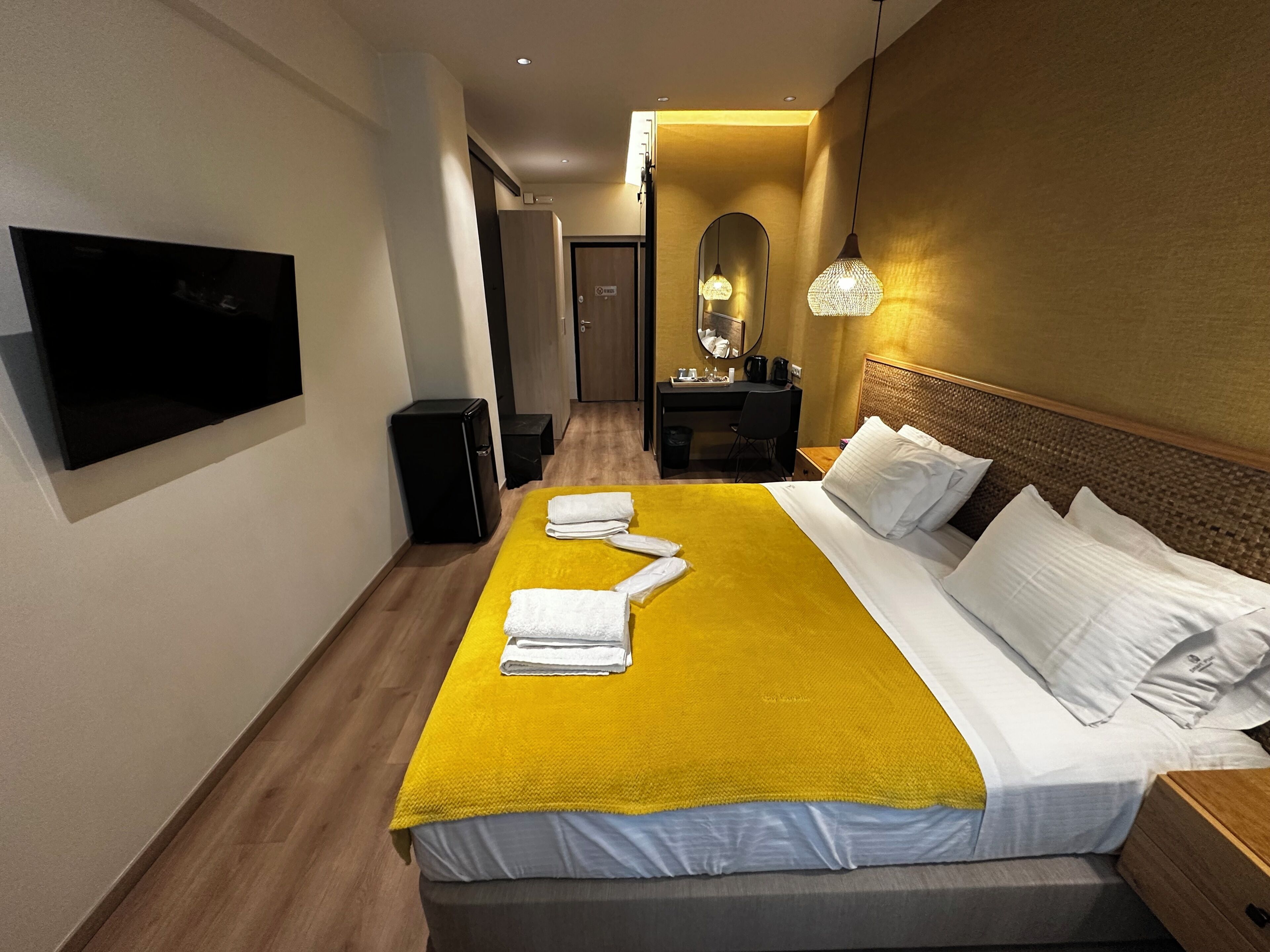 A hotel room with a bed, a flat-screen TV, a desk with a chair, a mirror, and a hanging light fixture.