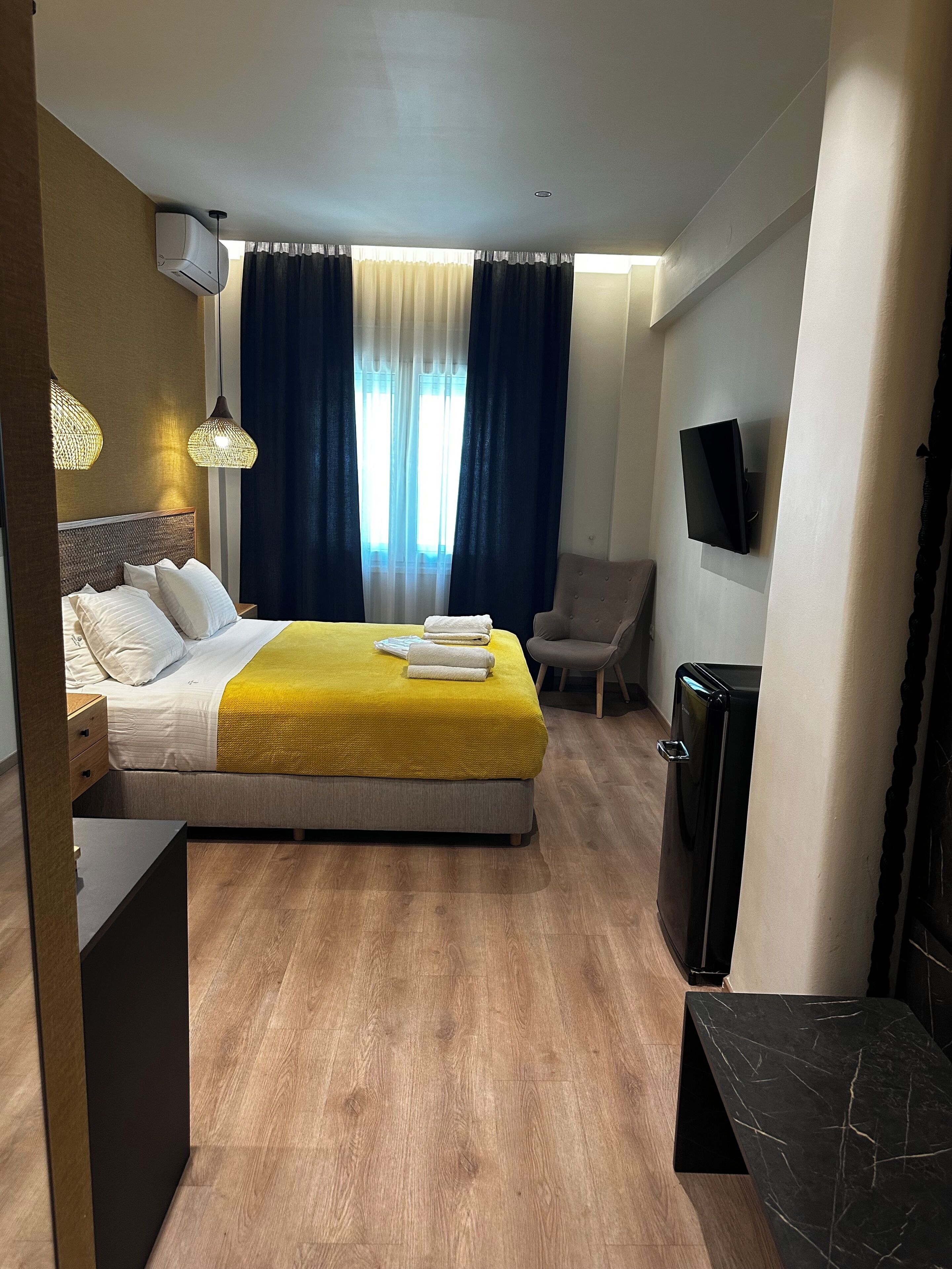 Habitació d'hotel amb llit, taula de nit, làmpada, cadira, televisió i un petit objecte negre a terra.