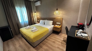 A hotel room with a bed, bedside table, desk, chair, and an air conditioning unit. - Syntagma Divine Guestrooms (Athens)