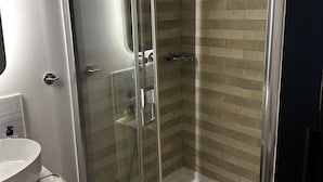 A glass-enclosed shower with a tiled wall, a white sink, and a mirror. - Syntagma Divine Guestrooms (Athens)