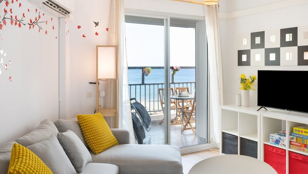 Una sala de estar con sofá, cojines amarillos, televisor y balcón con vistas.