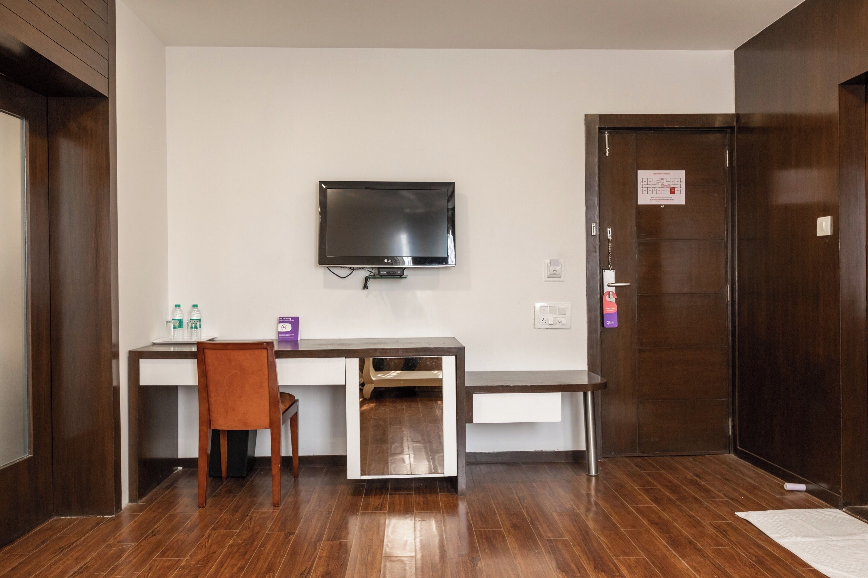 A hotel room with a wooden desk, a chair, a flat-screen TV mounted on the wall, and a door with a sign.