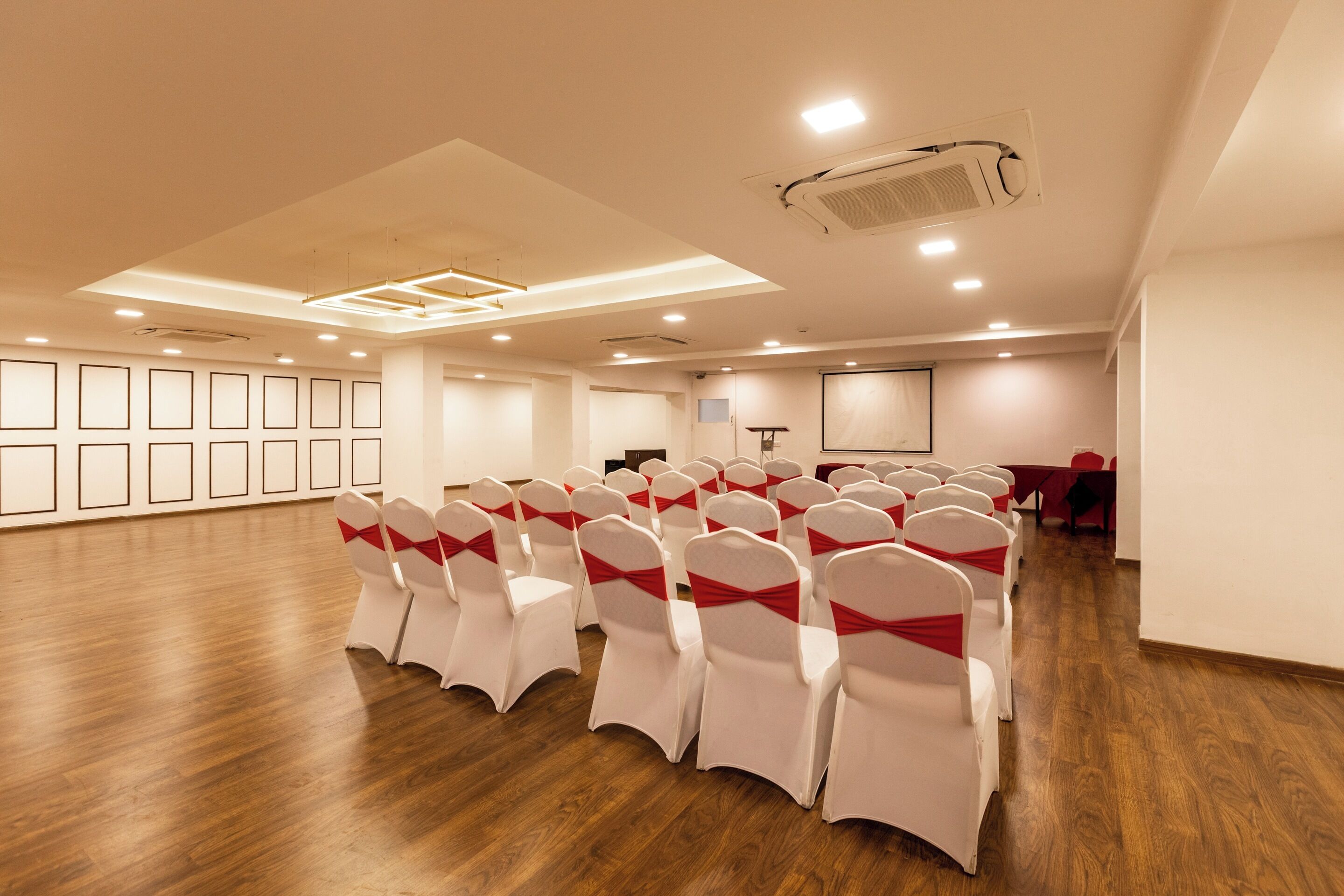 A conference room with rows of white chairs with red sashes, wooden flooring, recessed ceiling lights, and a projector screen.
