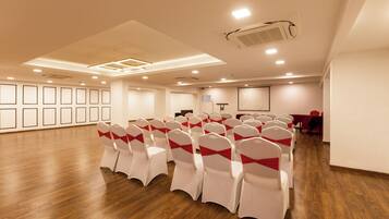 Un salón de conferencias con filas de sillas blancas con cintas rojas, suelo de madera, luces empotradas en el techo y una pantalla de proyector.