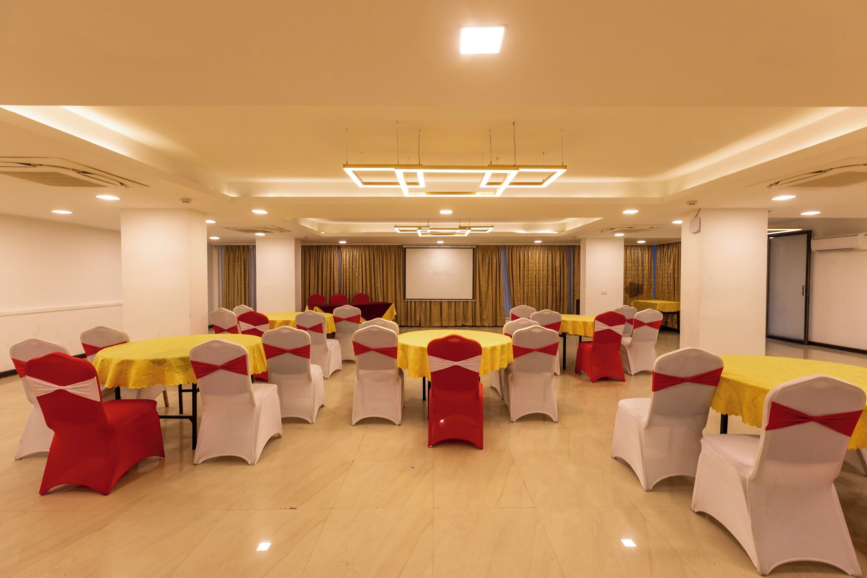 Une salle de conférence dotée de plusieurs tables et chaises, équipée de luminaires au plafond et d’un écran de projection.