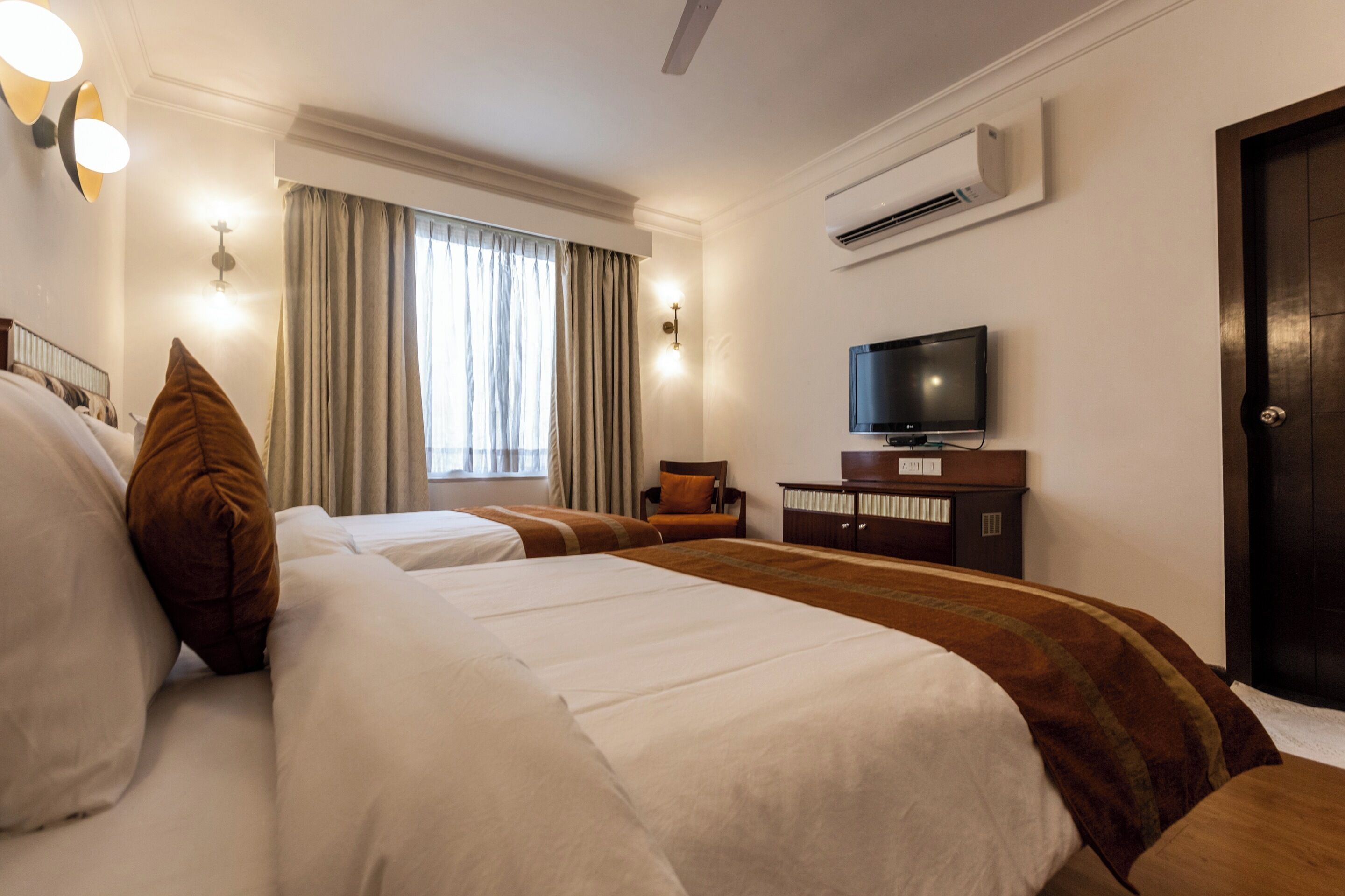 A hotel room with two beds, a television, a desk, and an air conditioning unit.