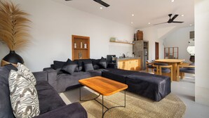 A modern living room with a sectional sofa, a wooden coffee table, ceiling fans, a kitchen area, and a dining table with chairs. - Villa Cinta Rose (Badung)