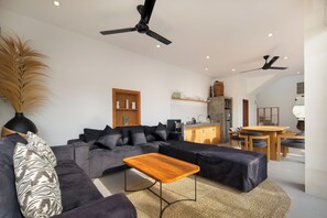 A modern living room with a sectional sofa, a wooden coffee table, ceiling fans, a kitchen area, and a dining table with chairs. - Villa Cinta Rose (Badung)