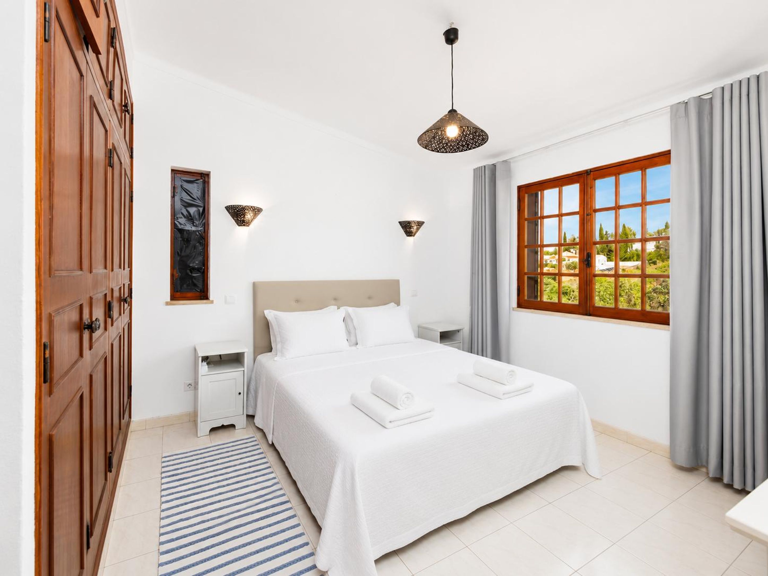 A hotel room with a bed, bedside tables, a window with curtains, a hanging light fixture, and a door with a wooden panel.