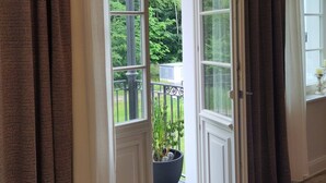 Une porte-fenêtre ouverte donne sur un balcon agrémenté d’un pot de fleurs, d’où l’on peut admirer la verdure, et dont le sol est en bois.