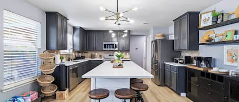Una cocina moderna con armarios oscuros, una isla central con taburetes, una lámpara de araña y un refrigerador.